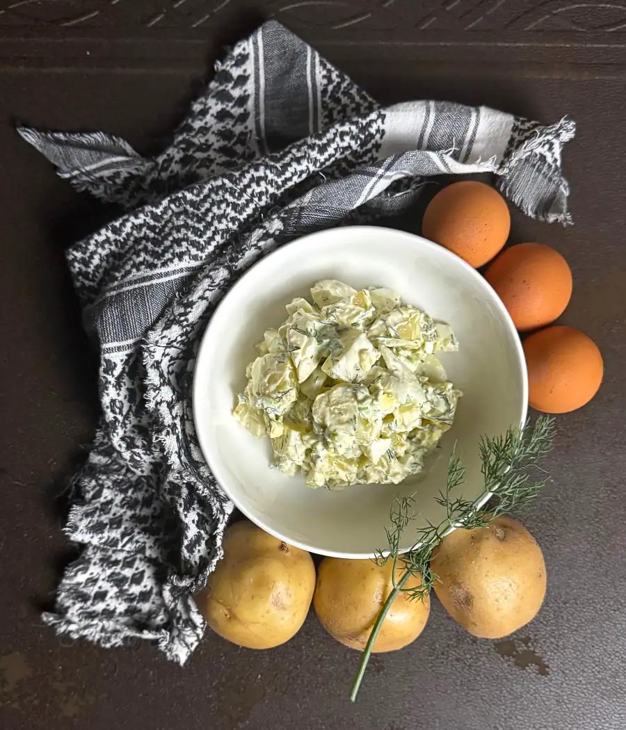 Bowl of creamy dill potato salad surrounded by eggs, baby potatoes, and fresh dill on a dark surface