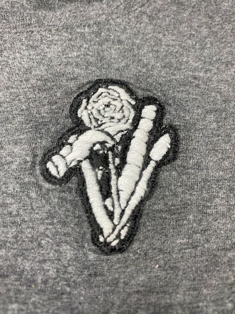Embroidered Craft Construct Create logo on a shirt dark grey shirt. A bouquet of a hammer, rose, ruler, and paint brush made from white thread with a black outline.