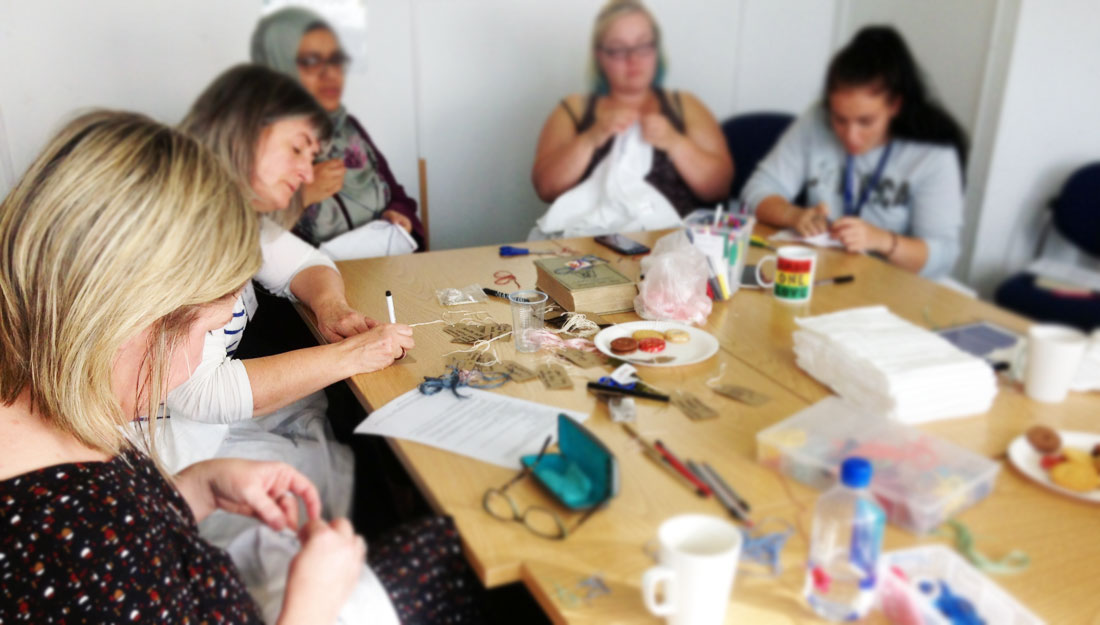 A group of 5 women sit around a table, concentrated. Some are embroidering baby muslins.