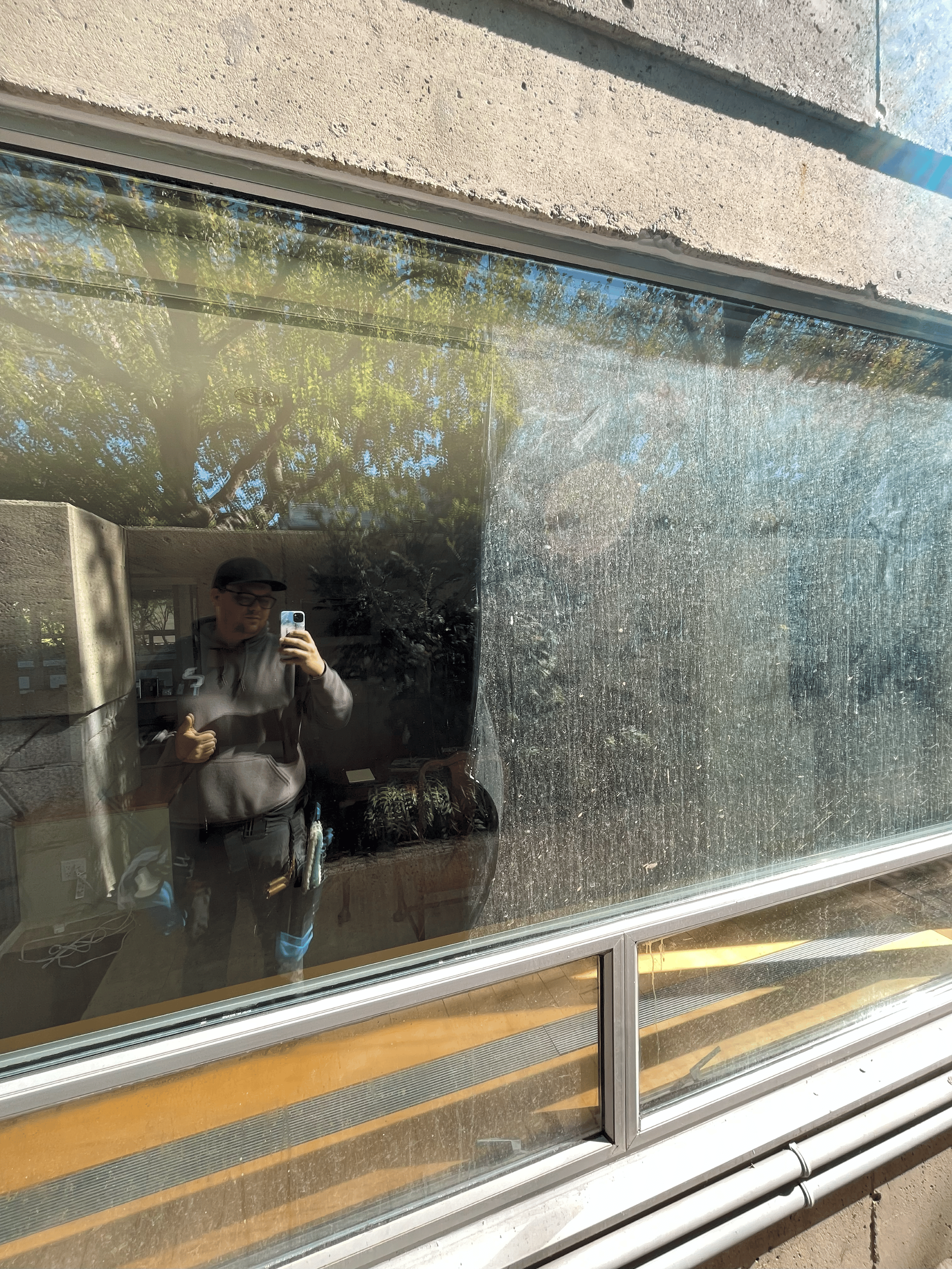 a craig and company contracting professional window cleaning technician stands in front of a large pane of glass taking a picture of themself in the reflection of the clean side while half of the glass remains dirty after a construction project, demonstrating the skill of the technician in post construction cleaning