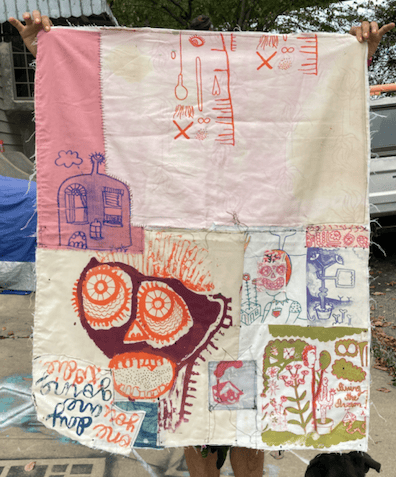 A textile piece composed of various pieces of fabric hand stitched together with irregular lines. Each piece of fabric is a slightly different muted color with a screen printed image on it in different colors, such as red, green, brown, and blue. Some of the screen printed images include a simple illustration of a bottle shaped house, a skeleton-reminiscent figure with artistically rendered large eyes and mouth, an upside-down cursive rendering of the words “some day you are going to die,” and a garden like scene with the words “living the dream.”