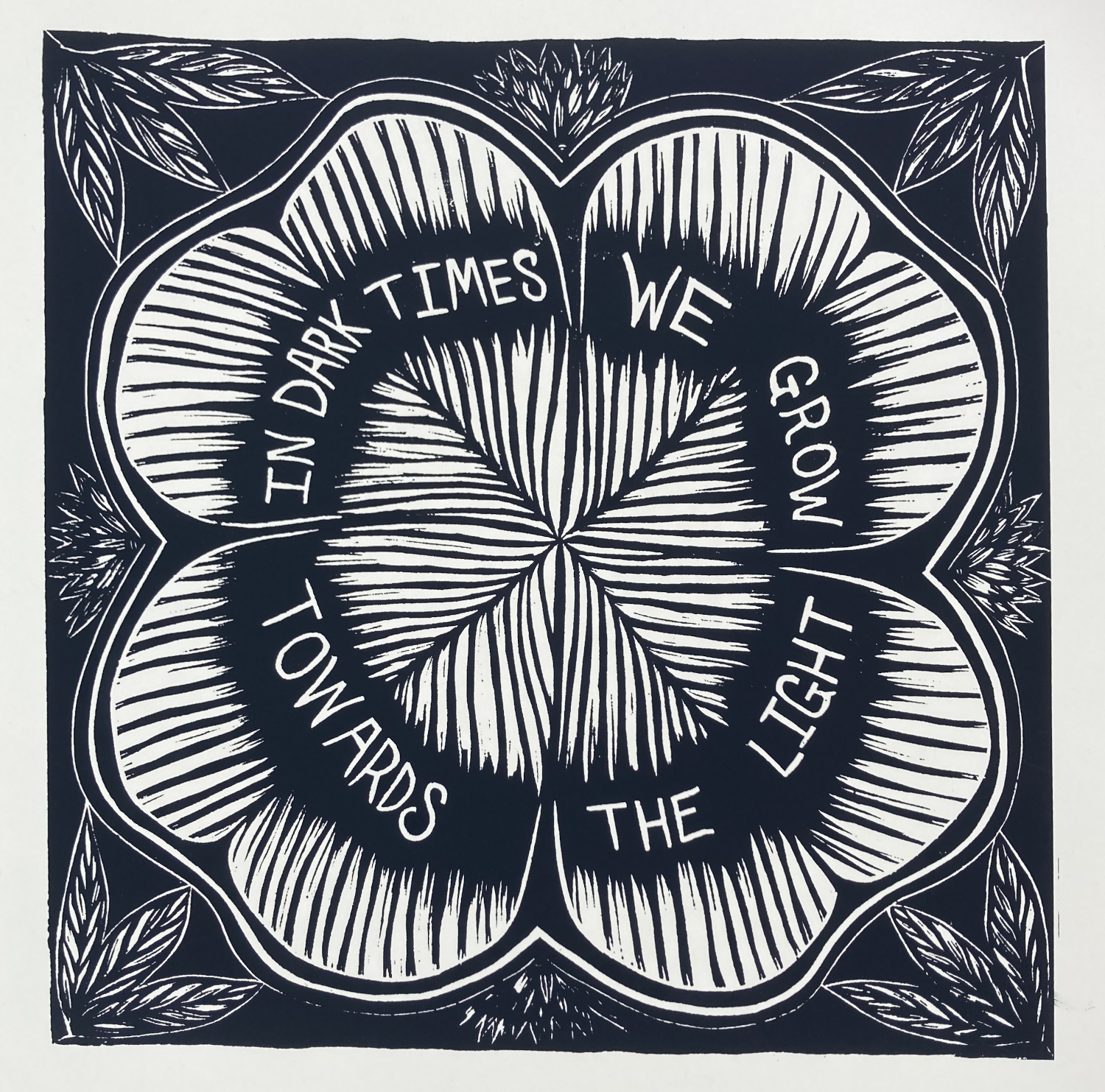 A screenprint of dark blueish black ink on cream colored paper, depicting a flower-like geometric shape within which appear the words "In dark times we grow towards the light"