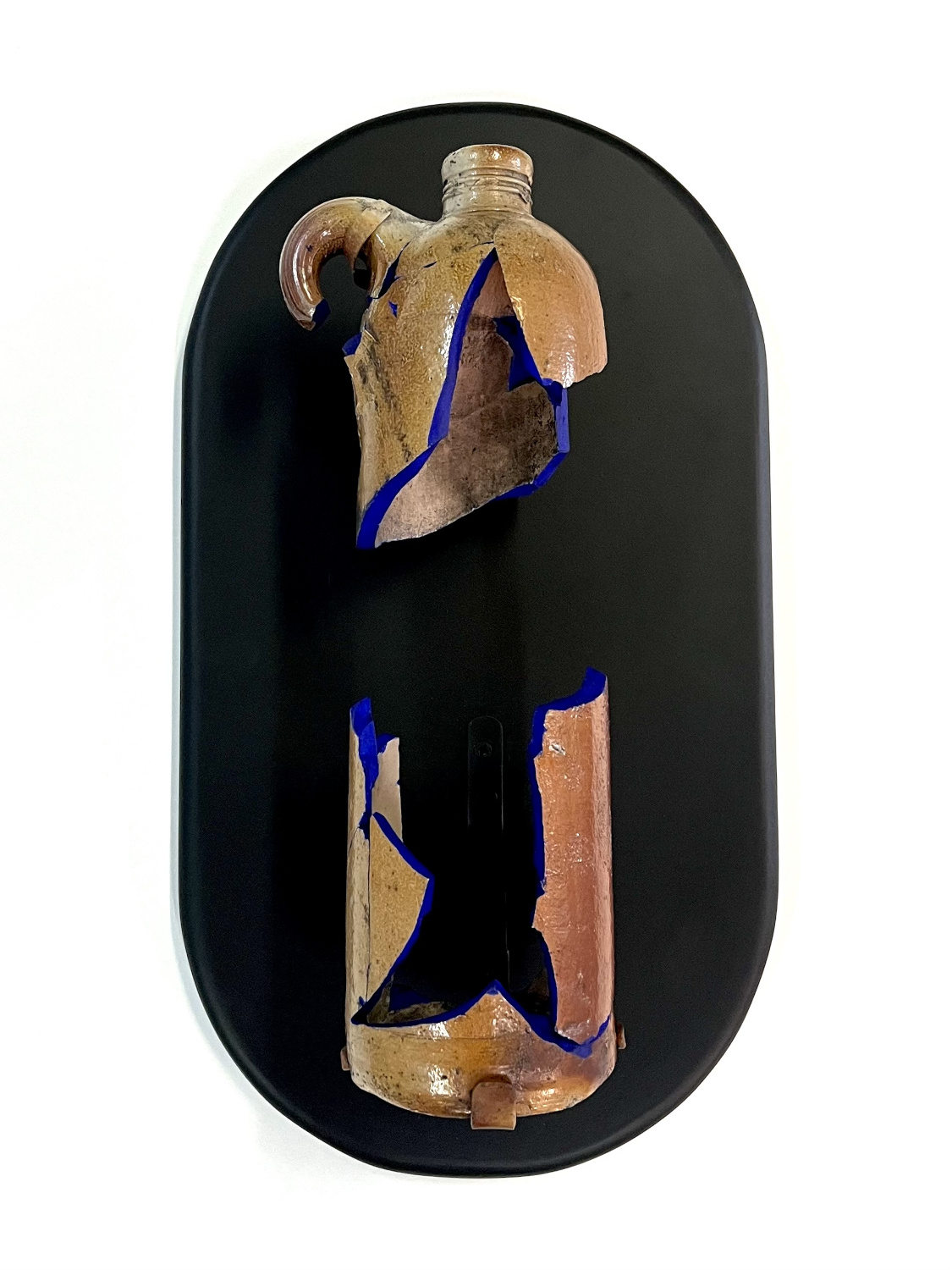 A sculpture of a reconstructed 1800’s ceramic bottle which remains broken in half with its two pieces set in vertical orientation without touching, standing at 12” tall and floating on a black oval wall mount. The bottle’s exterior surface is a high gloss glaze finish of variable earthen tones, the interior has is matte, shades of beige with signs of dirt still evident. The bottle’s broken edges are painted an ultramarine blue, separating the exterior and interior surfaces of the bottle.