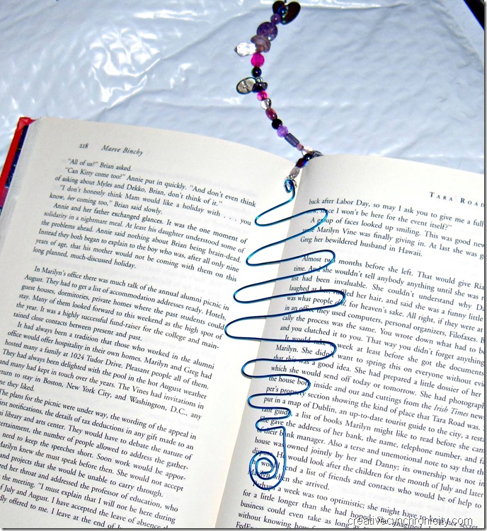 Wire and bead bookmarks CreativeCynchronicity.com the finished bookmark with blue rounded zigzag wire and assorted beads in pinks and purples. The wire part is sitting in an open book to demonstrate how these work.