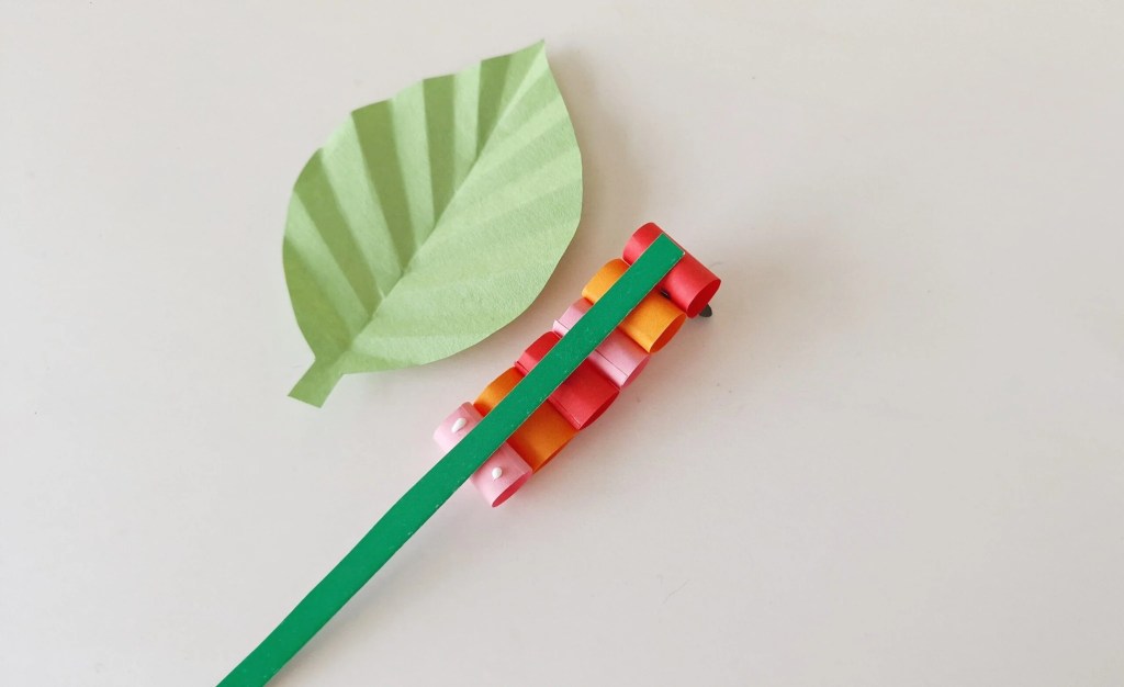 Green paper strip threaded through the paper caterpillar beside the folded leaf.
