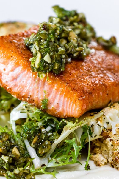 A seared salmon fillet topped with green relish, served over leafy greens, cauliflower, and creamy sauce on a plate.