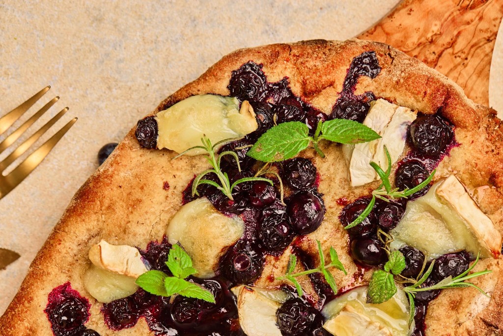 Close-up of baked blueberry goat cheese flatbread with melted goat cheese, roasted blueberries, rosemary, and fresh mint.