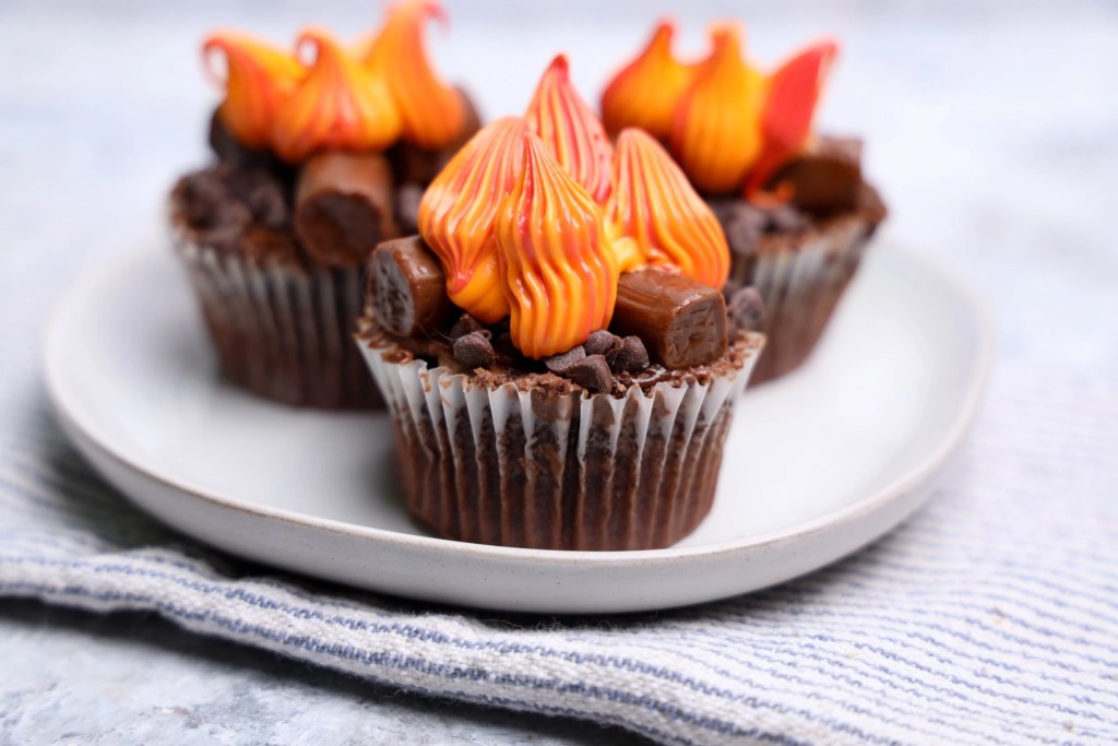 Close-up of a campfire cupcake decorated with Tootsie Roll logs, chocolate chips, and colorful piped frosting flames.