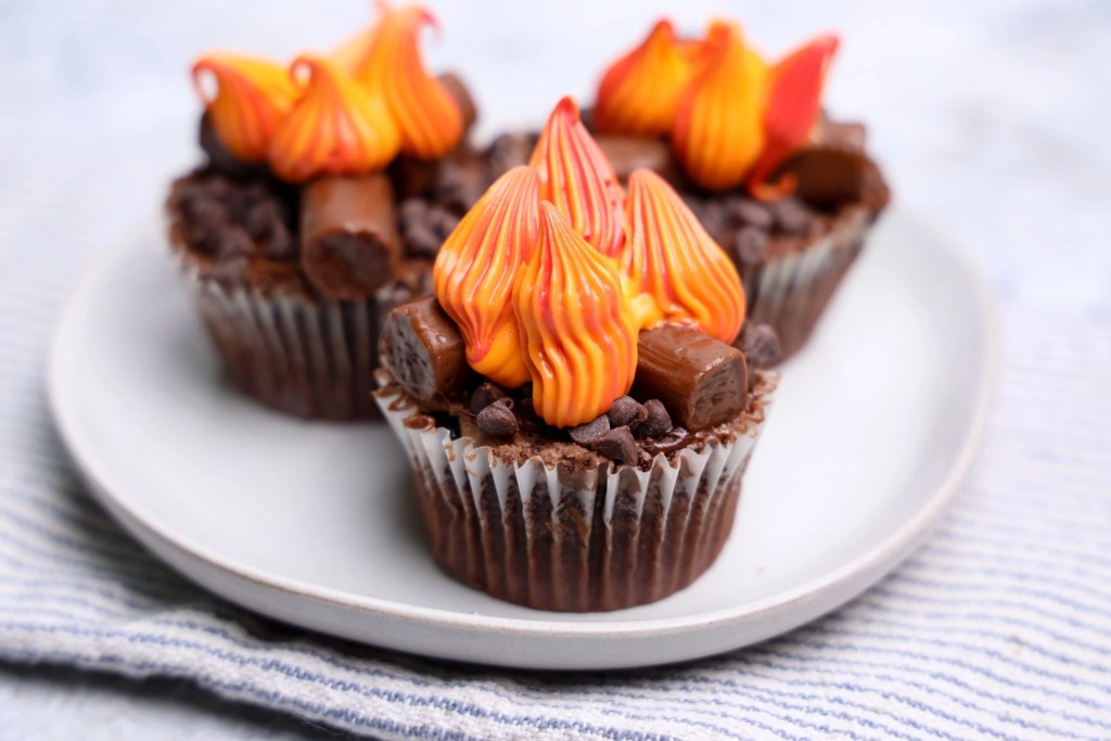 Three finished campfire cupcakes decorated with candy logs, chocolate chips, and colorful frosting flames.