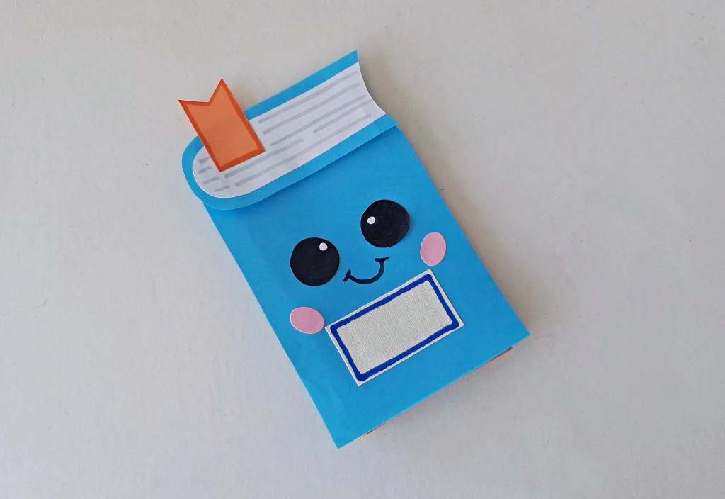 Finished blue notebook paper bag puppet with a smiling face and orange bookmark.