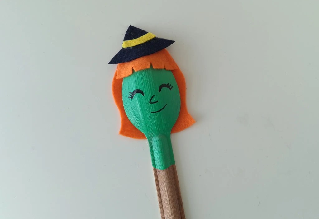 Painted green spoon witch with a smiling face, orange hair, and felt hat.