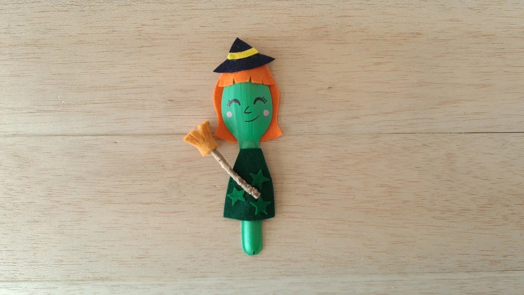 Finished wooden spoon witch with orange felt hair, a green dress, and a miniature broom.