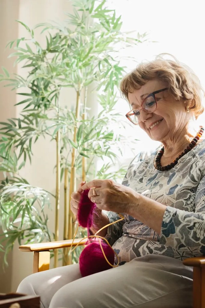 Older woman smiling as she knits with magenta yarn in a sunny room.