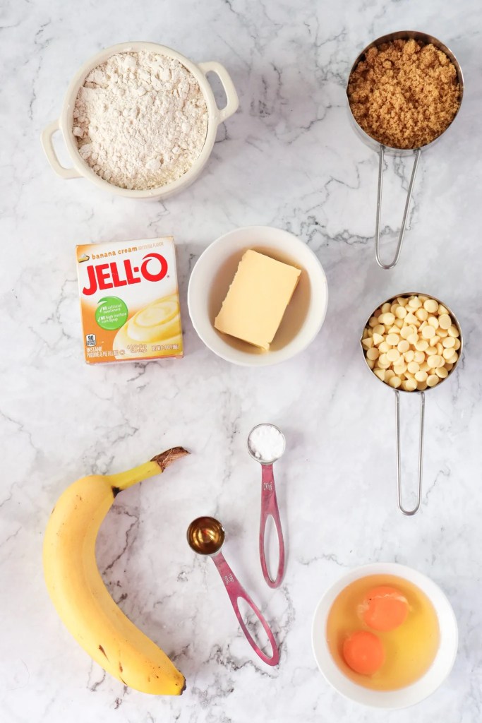 Ingredients for banana pudding cookies including banana, pudding mix, flour, brown sugar, butter, eggs, and white chocolate chips