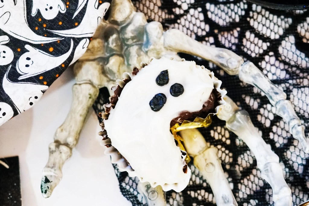 Close-up of a chocolate ghost cupcake decorated with white frosting and three black icing dots for a spooky face.