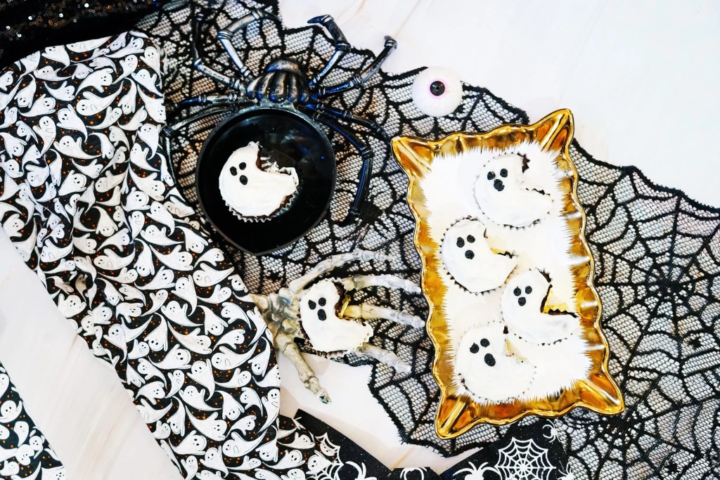 Ghost-shaped chocolate cupcakes with white frosting and black icing faces arranged on a gold tray with Halloween decorations.