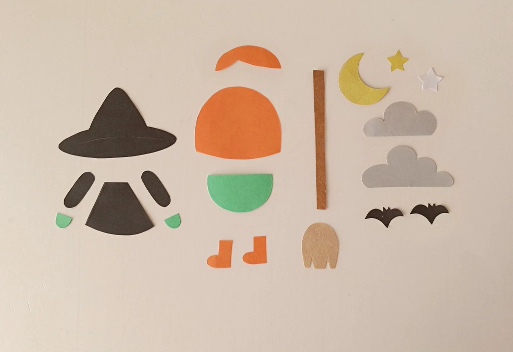 Colored paper cutouts for a flying witch Halloween craft, including a black witch hat, orange hair, green hands, broomstick, moon, stars, and bats.