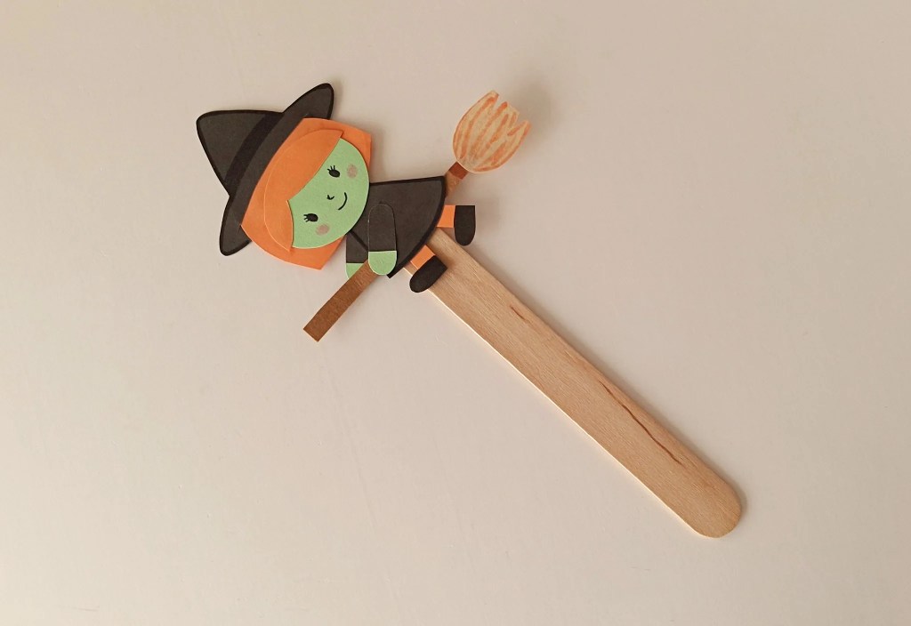 Handmade paper witch riding a broomstick attached to a wooden popsicle stick for an interactive Halloween craft.