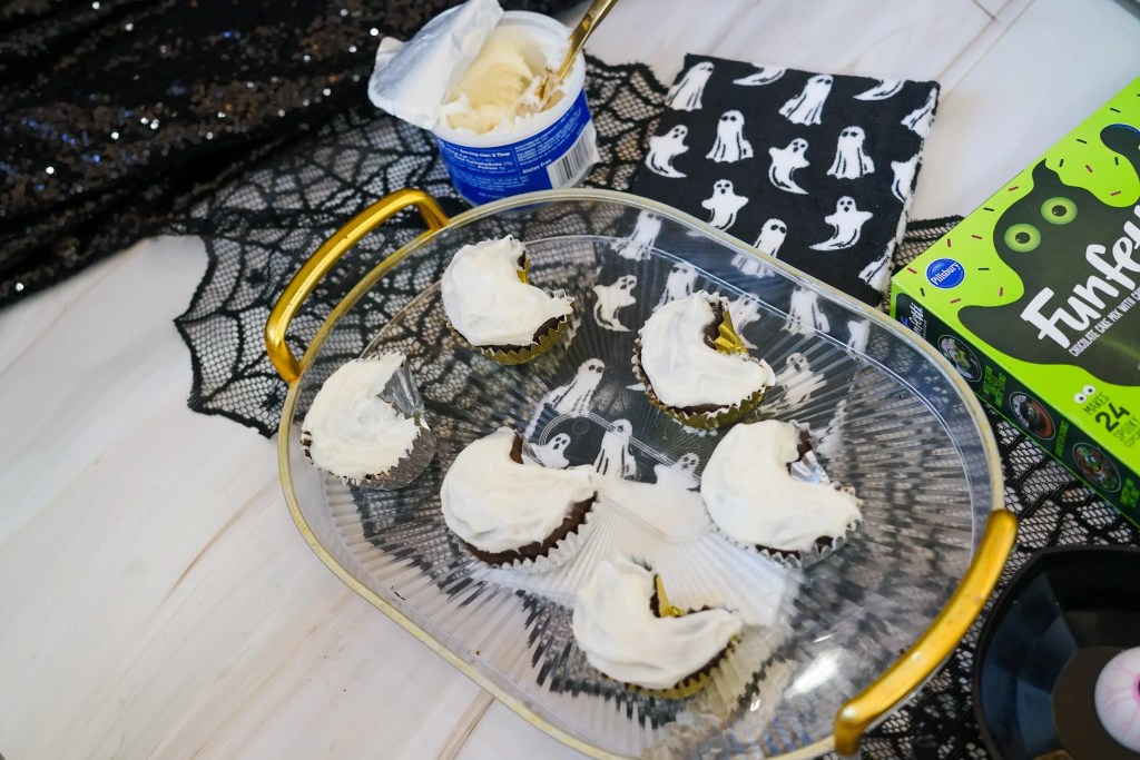 Ghost-shaped chocolate cupcakes covered with white vanilla frosting before their faces are added.