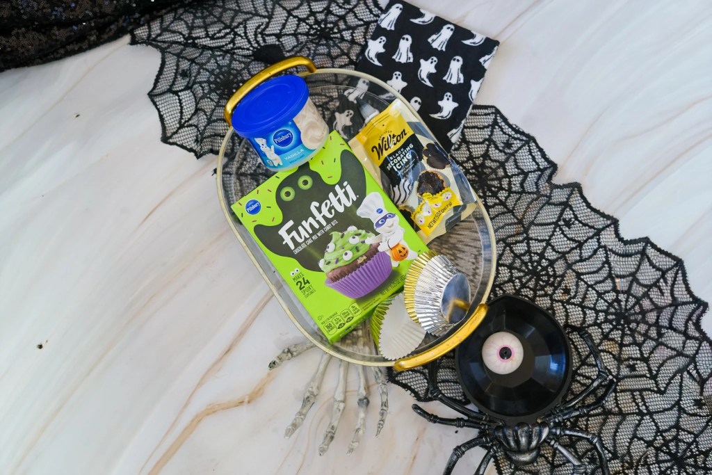 Chocolate cake mix, vanilla frosting, black decorating icing, and cupcake liners arranged for making Halloween ghost cupcakes.