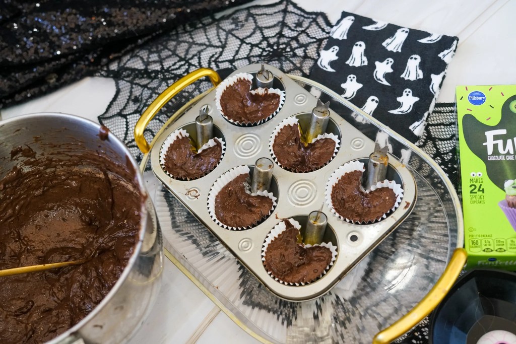 Chocolate cupcake batter in paper liners shaped into ghosts using metal holders positioned inside a muffin pan.