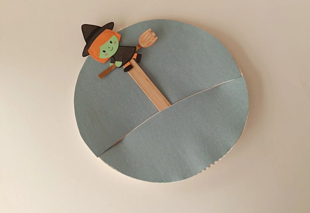 Paper witch attached to a popsicle stick being inserted through the slit in a blue paper plate Halloween scene.