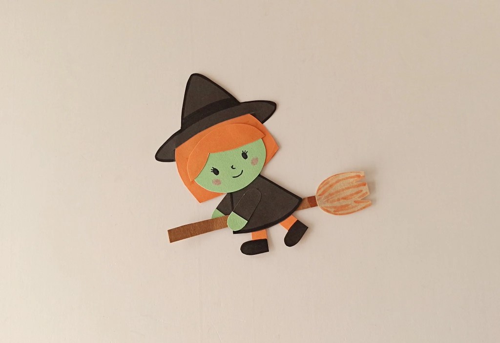 Smiling paper witch with green face, orange hair, and black hat riding a handmade paper broomstick.