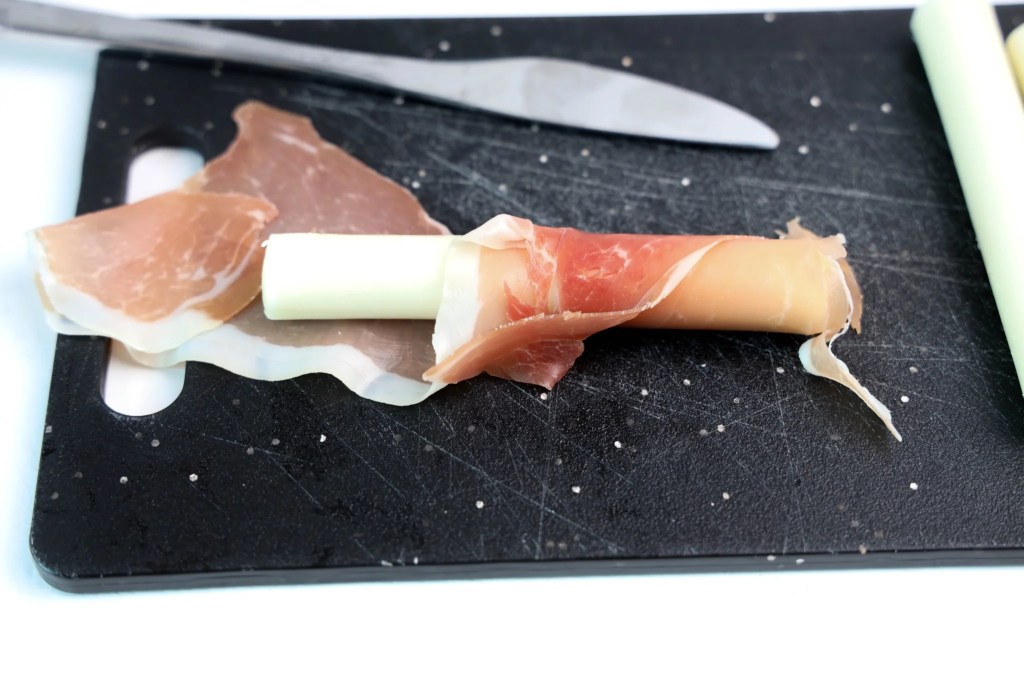 Mozzarella string cheese being wrapped in a slice of prosciutto on a cutting board.