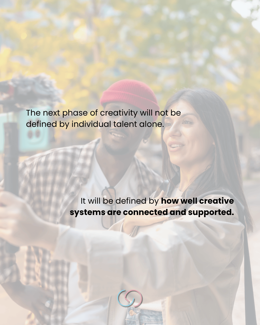 A woman and a man engaged in a conversation, with the woman holding a smartphone as part of a creative collaboration. Background features blurred trees with autumn leaves.