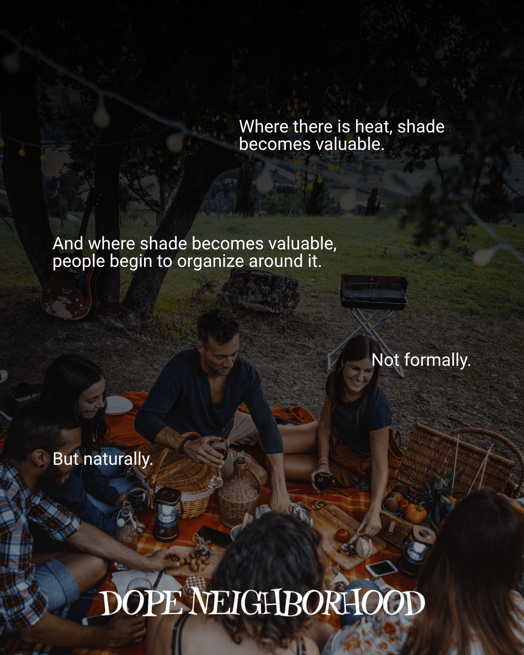 A group of people sitting on a picnic blanket under the shade of trees, enjoying food and drinks together with a guitar and a small grill in the background.