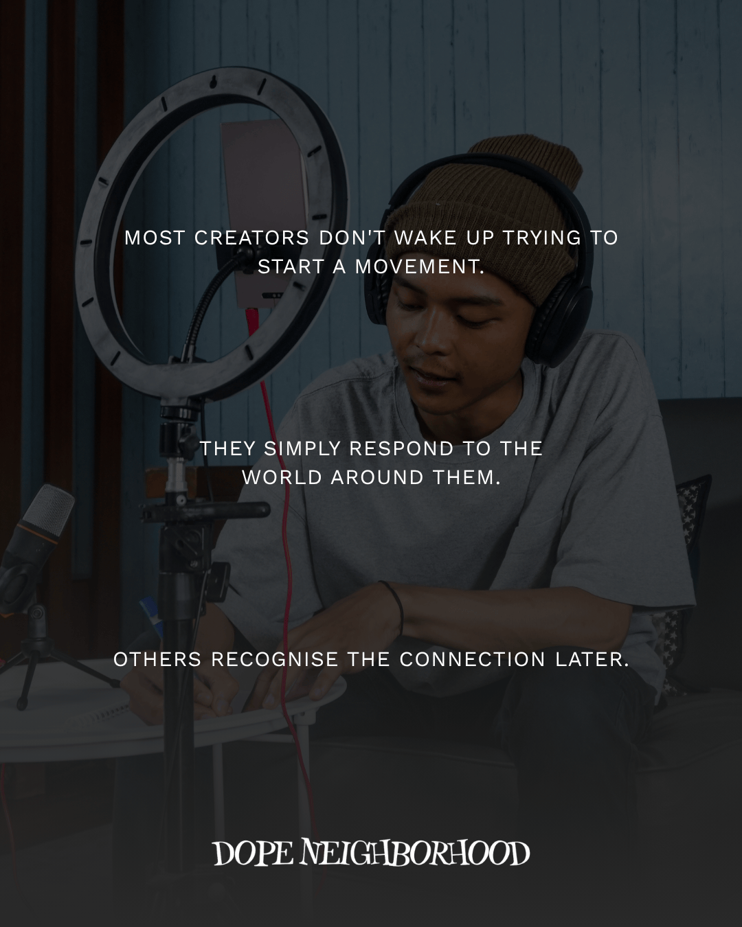 A young man wearing headphones and a beanie is sitting at a table with a microphone and a ring light, engaged in a creative process.