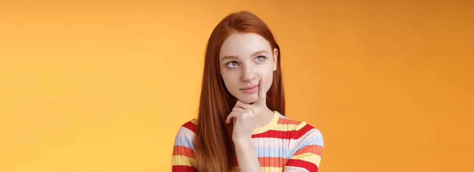 A young woman with red hair thinking about Real User Monitoring, touching her chin, against an orange background.