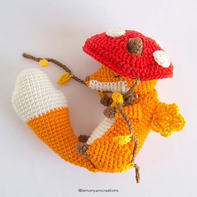 un renard réalisé au crochet qui tient une branche dans sa bouche, avec des pommes de pin