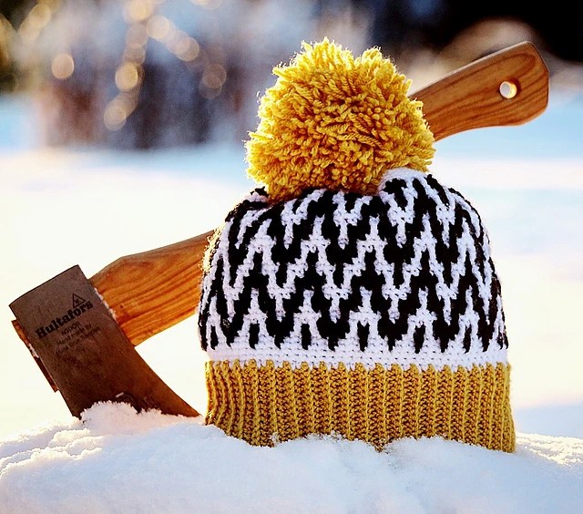 bonnet jaune blanc et noir, avec pompon jaune dans la neige avec une hache réalisé en crochet mosaique