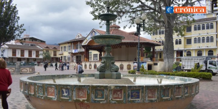 A historic plaza featuring a decorative fountain at the center, with trees and buildings in the background under a cloudy sky.