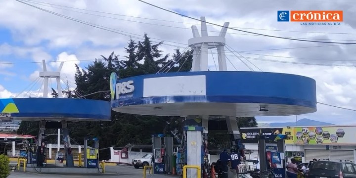Gas station with two fuel pumps under a blue canopy, surrounded by trees and a clear sky.