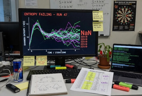 Desk workspace with a monitor showing an entropy plot labeled 'Entropy Failing - Run 47' and multiple colored lines, plus a laptop, notebook, markers, and a plant on the desk.