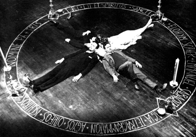Three people lying in a circular formation on a wooden floor, surrounded by a mystical symbol and candles.