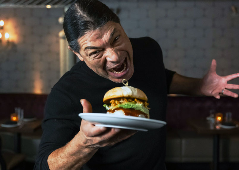 A man in a black shirt holding a plate with a large cheeseburger, gesturing excitedly toward the camera in a dim restaurant.