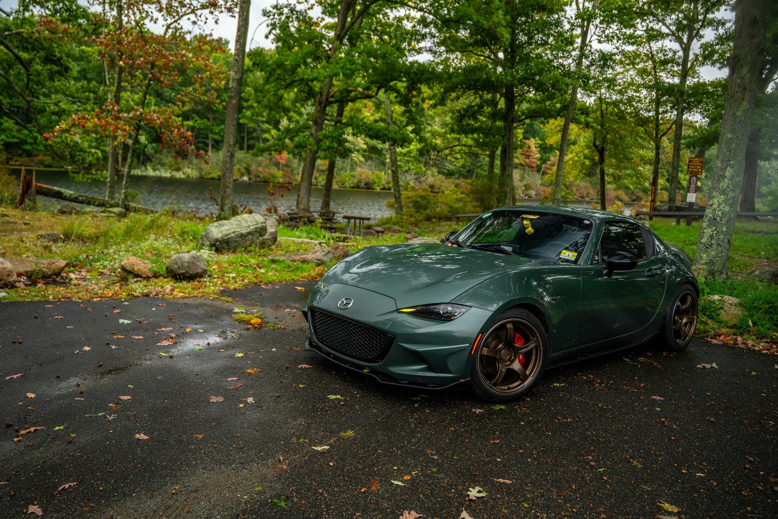 Green Mazda Miata RF on bronze wheels parked by a lake in the New York countryside with fall trees