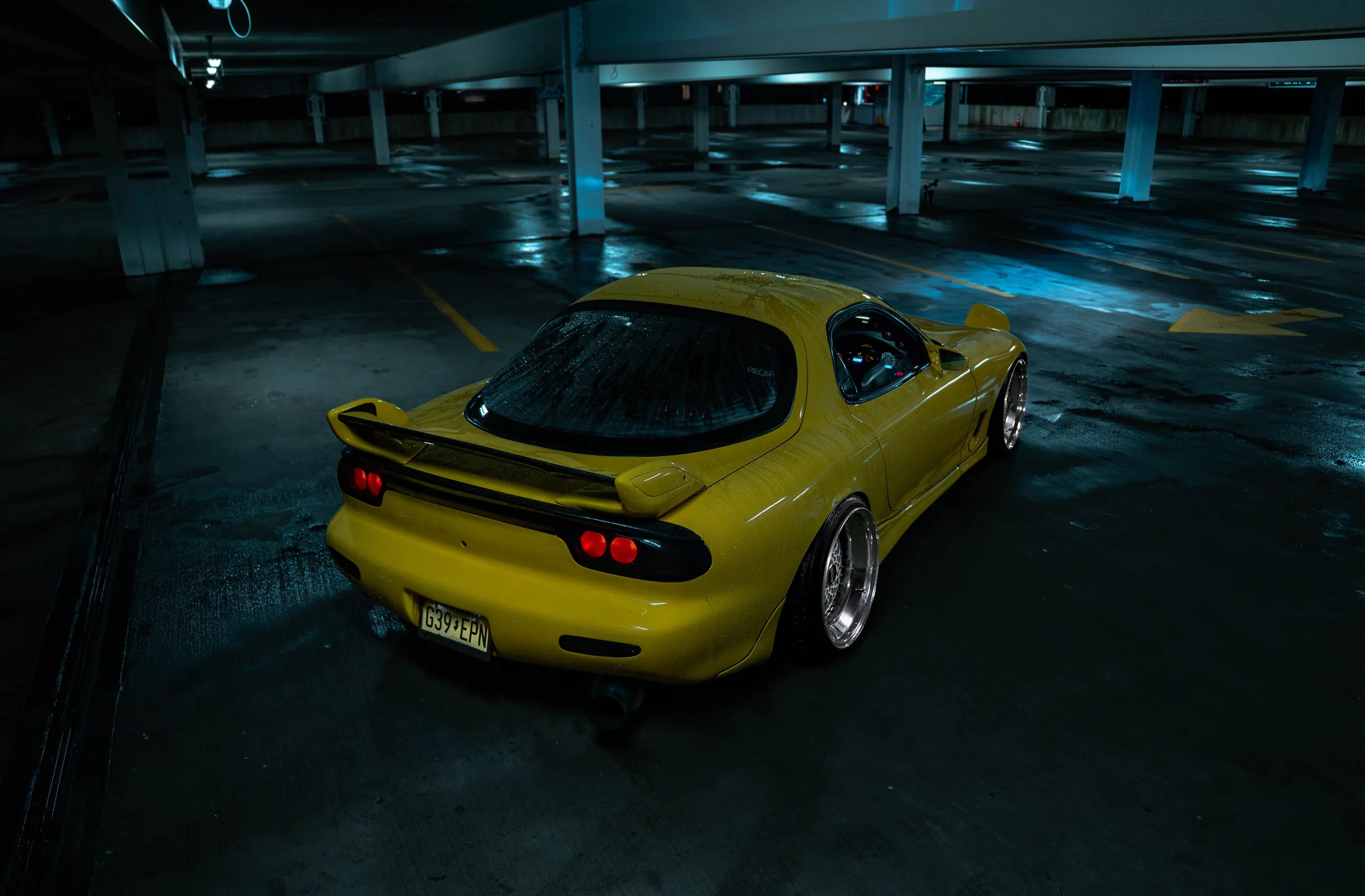 Rear three-quarter view of a yellow widebody Mazda RX7 FD with a rear wing on a wet garage floor