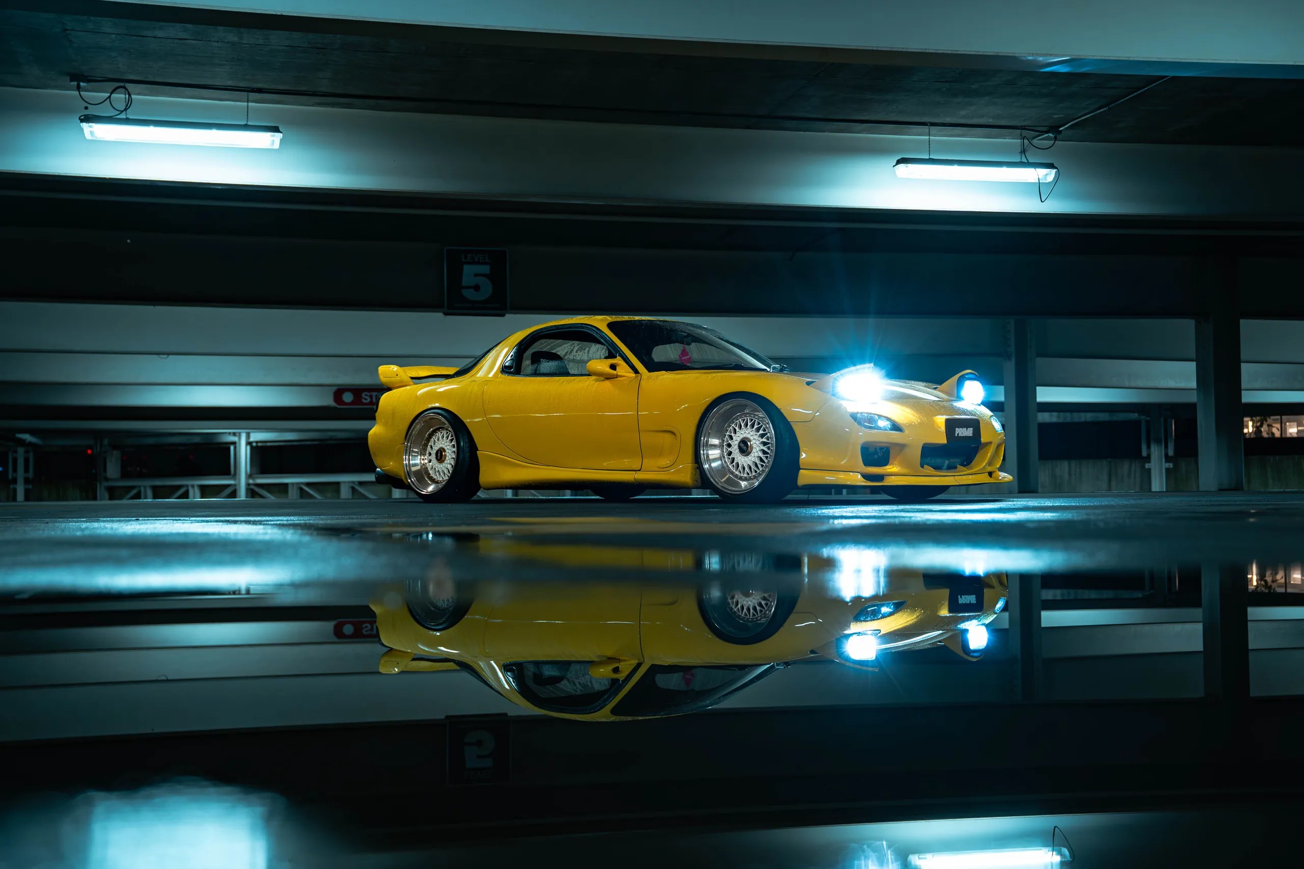 Yellow Mazda RX7 FD with headlights up, reflected in a wet parking garage floor