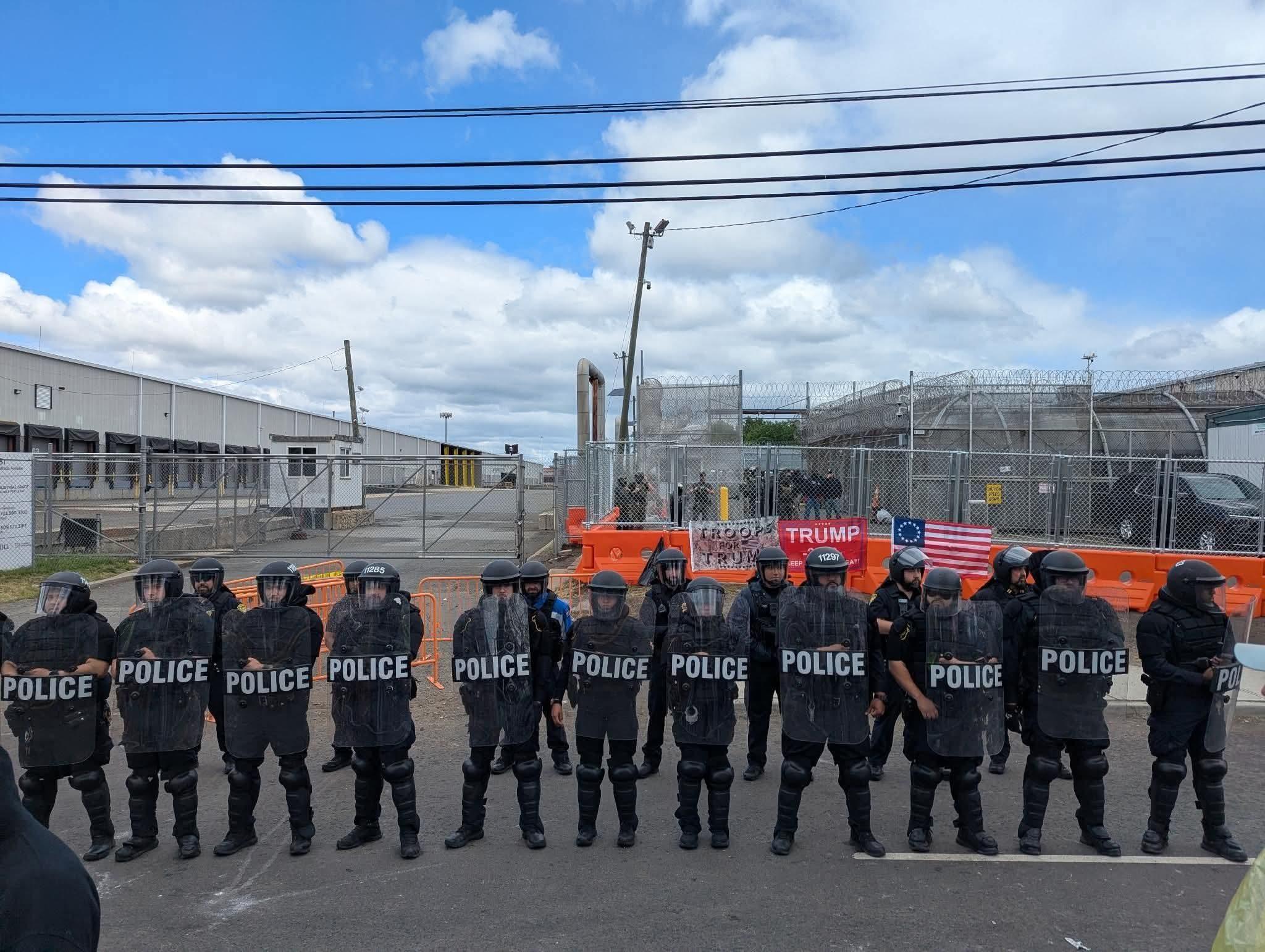 17 police agents with riot shields standin in a row in front of a barrier with pro trump flags