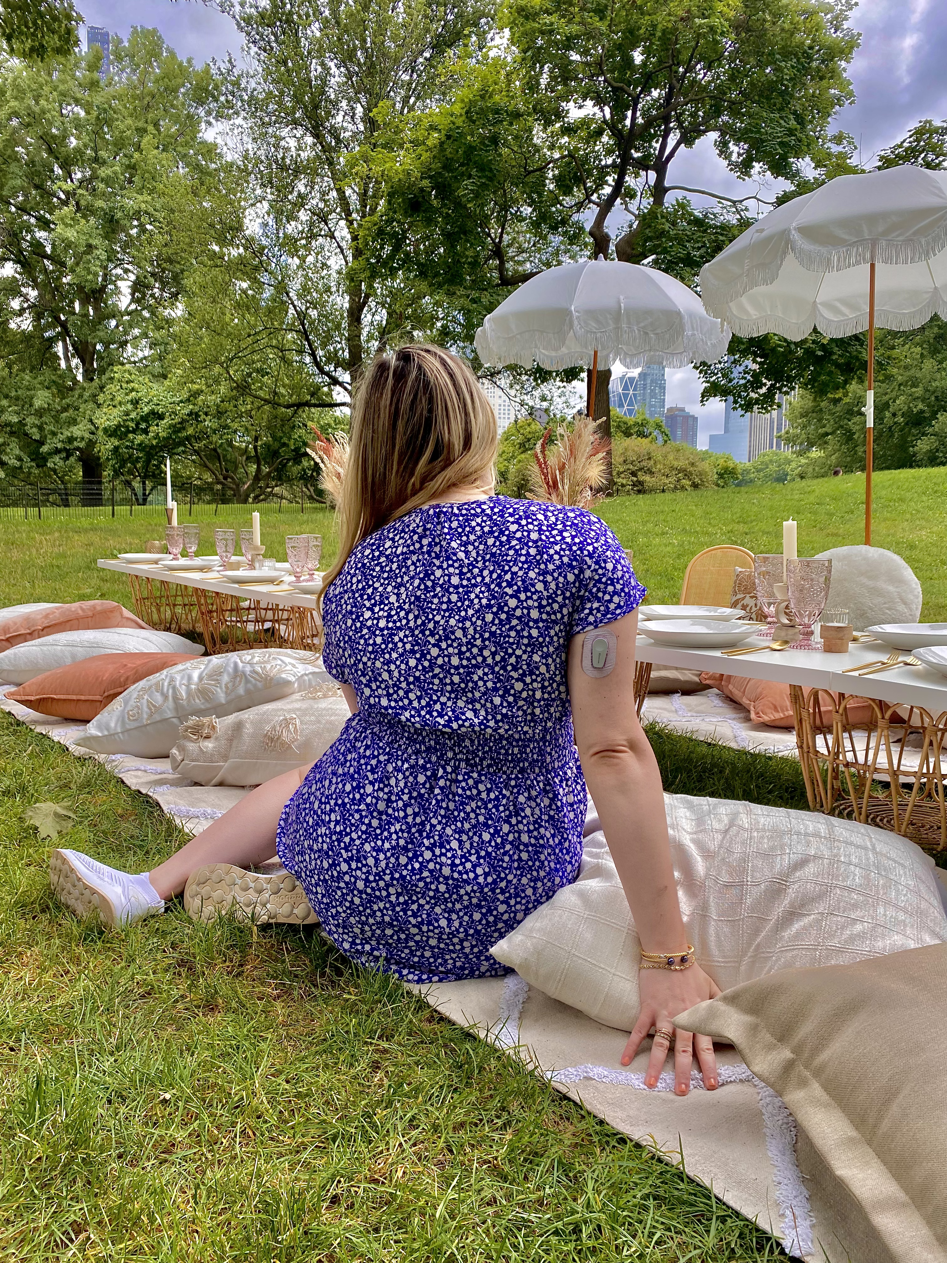 Woman in a blue patterned dress sitting on a pillow for a picnic in Central Park. She is facing away and has a dexcom on her right arm.