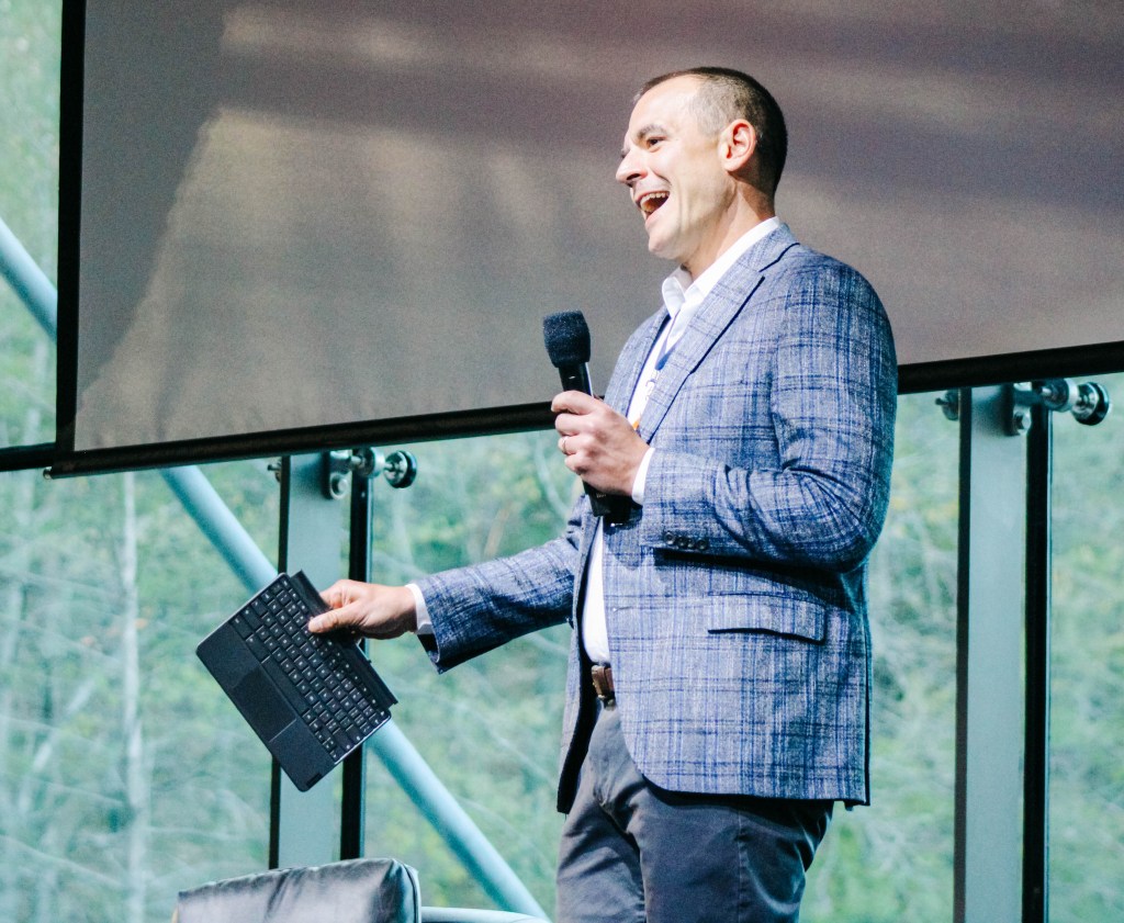 A speaker holding a microphone and a laptop while presenting on stage at a conference.