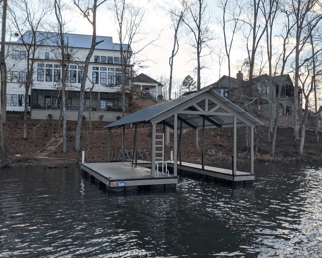 Custom dock with a pitched roof, overlooking a lakeside home and surrounding trees during the winter season.