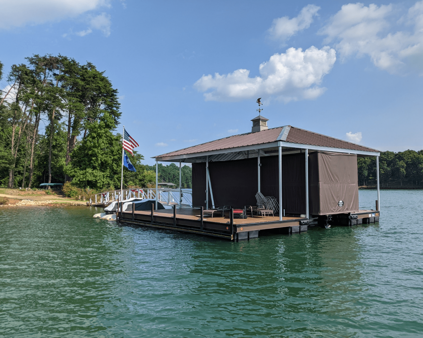 Floating covered dock on a lake with a Touchless Boat Cover system, patio seating, and American flags, surrounded by clear water and trees under a sunny sky.