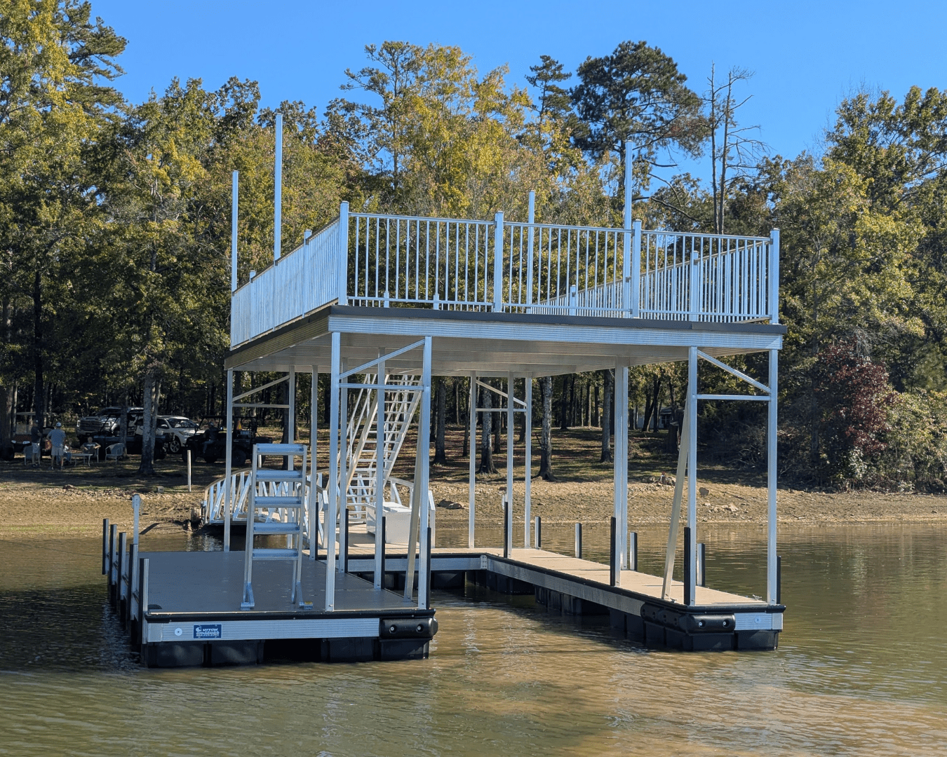 Two-level aluminum boat dock with a rooftop deck, white railing, and floating platforms on a calm lake, surrounded by trees and shoreline.
