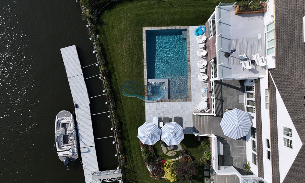 rectangular shaped inground pool and patio with umbrellas and white chairs in house by shore, boat docked in water