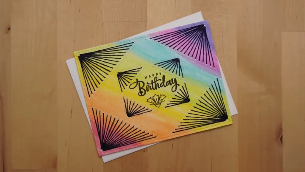A colorful handmade birthday card on a wooden table, featuring "Happy Birthday" in the center, with geometric black fan patterns on a gradient background of yellow, green, blue, and pink hues. The card also showcases a small stitched rainbow design in one corner for added charm.